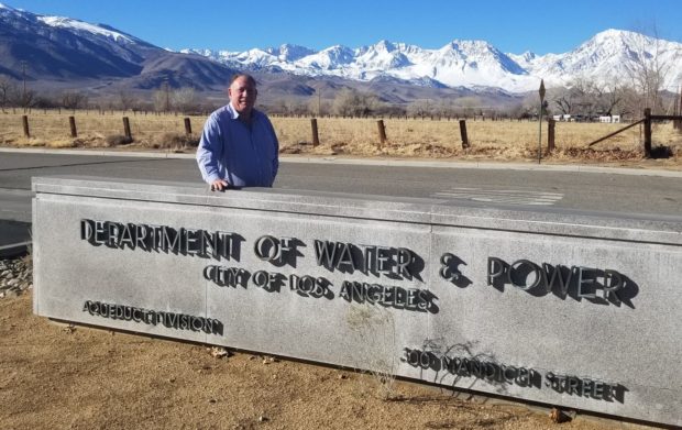 man standing behind Los Angeles Department of Water and Power sign with snow capped mountains.